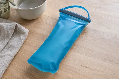 Réservoir d’hydratation souple bleu Malibu avec fermeture coulissante et poignée flexible, posé sur du bois clair.
