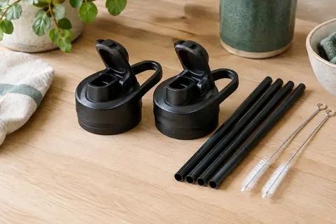 Deux bouchons noirs à paille accompagnés de pailles de rechange et de brosses sur une table claire.