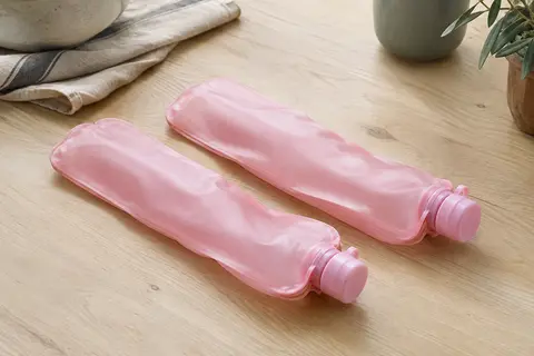 Deux flasques souples roses posées sur une table en bois clair près d’un linge en lin naturel.