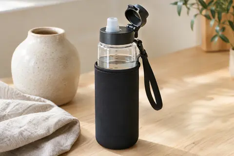 Gourde Reeho en verre avec housse noire, couvercle rabattable et paille posée sur une table claire.